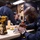 Three individuals wearing matching navy blue uniforms are working at a woodworking workbench in what appears to be a military workshop or carpentry shop. The foreground subject, wearing safety glasses, is carefully marking or measuring a piece of wood with a pencil and square. The workspace is equipped with power tools and woodworking supplies.