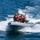 A gray rigid inflatable boat (RIB) is moving at high speed across open blue water, creating significant wake and spray. Four individuals are aboard, wearing orange life vests and at least one yellow hard hat, with a small American flag visible at the stern. The scene appears to be a military or coast guard operation based on the vessel type, gray color scheme, and personnel gear.