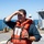 A man wearing an orange life vest stands on the deck of a vessel, shielding his eyes from the sun while looking into the distance. He holds a small red object in his other hand. A second crew member wearing a white hard hat is visible in the background near gray metal deck equipment.