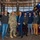 A group of approximately 12 men pose for a group photo inside what appears to be a space or aerospace facility. One individual is wearing a U.S. Army military uniform in camouflage pattern, while others are dressed in business casual or casual attire. Behind the group is a large display model of a satellite with blue solar panels.