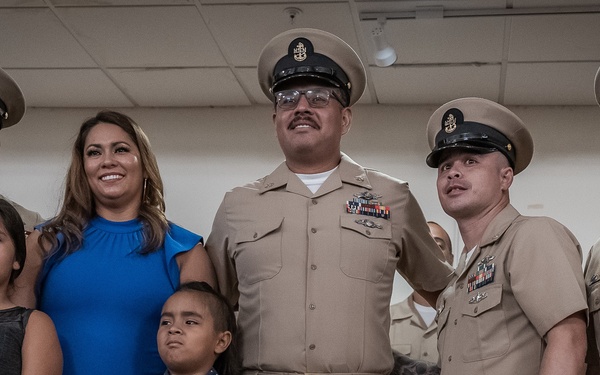 A group photo taken at what appears to be a U.S. Navy chief petty officer pinning or promotion ceremony. Four uniformed Navy personnel wearing khaki service uniforms with chief petty officer covers (hats) are posing alongside a woman in a blue dress and two children. The setting is an indoor venue with drop ceiling tiles, consistent with a military community or conference room.