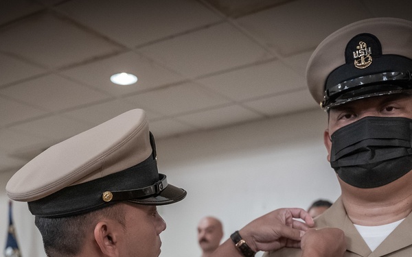 A U.S. Navy military ceremony is taking place in which one sailor is pinning an insignia or device onto another sailor's uniform collar. The recipient is wearing a Chief Petty Officer combination cover with the USN anchor device and a black face mask, along with multiple service ribbons and warfare badges. Several other uniformed personnel are visible in the background observing the ceremony.