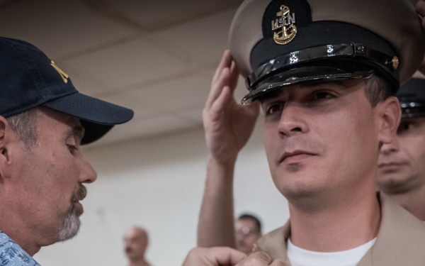 A U.S. Navy Chief Petty Officer promotion ceremony is taking place, with a civilian (likely a family member) pinning anchors or insignia onto the collar of the newly promoted sailor. Simultaneously, another person is placing the distinctive CPO combination cover (hat) on the inductee's head. This is consistent with the traditional U.S. Navy Chief Petty Officer pinning ceremony.
