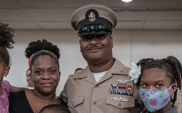 A U.S. Navy senior enlisted sailor, wearing a khaki service uniform with multiple ribbons and warfare devices, poses with his family during what appears to be a military promotion or recognition ceremony. He is surrounded by his wife and three children in a formal indoor setting. Other uniformed Navy personnel are visible standing in formation in the background.