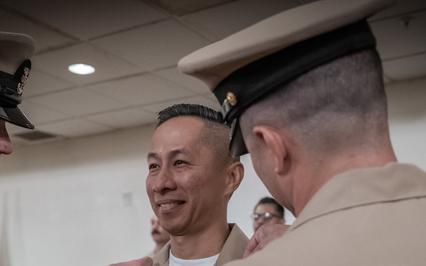 A military ceremony is taking place in an indoor setting where two uniformed naval officers are pinning insignia or a device onto a third service member's collar or uniform. The central figure is smiling, suggesting this is a positive recognition or promotion event. All individuals are wearing U.S. Navy service khaki uniforms.