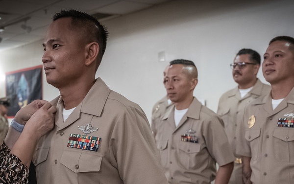 A military promotion or pinning ceremony is taking place, with a civilian woman pinning insignia or a collar device onto a U.S. Navy Chief Petty Officer's uniform. Several other Navy personnel in tan/khaki service uniforms stand in formation in the background. The setting appears to be an indoor military facility or event hall.