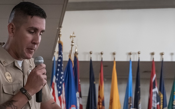 A U.S. Navy enlisted service member in tan/khaki uniform is speaking at a podium with a microphone during what appears to be a formal military ceremony. A white officer's cover (cap) rests on the podium, and a row of U.S. state and military flags is displayed in the background. Several other uniformed Navy personnel are seated in the background as an audience.