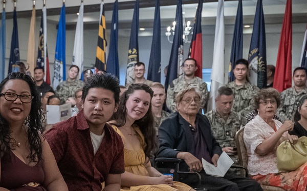 A formal military ceremony is taking place in an indoor venue, with civilians seated in the foreground as an audience and uniformed military personnel standing in the background. Multiple U.S. state and military flags are displayed along the back wall, suggesting an official event such as a promotion, retirement, or award ceremony. The attendees appear to be family members and guests of the service members present.