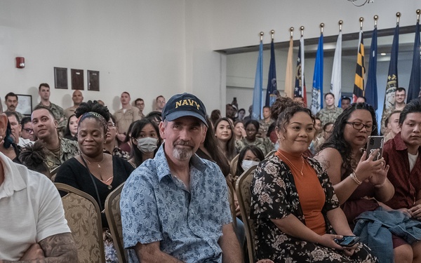 A large group of civilians and military personnel are gathered in a formal hall for what appears to be a military ceremony or event. The audience is seated in rows of gold chairs, with multiple state and military flags displayed prominently in the background. The atmosphere appears celebratory and relaxed, with attendees smiling and engaging with the event.