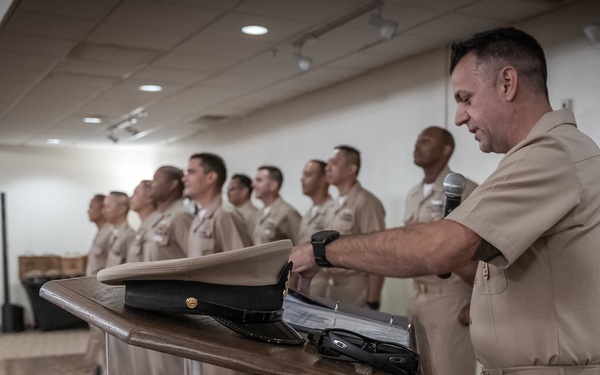 A military ceremony is taking place indoors, likely a U.S. Navy event, with personnel in tan/khaki service uniforms standing at attention in a formation. A senior enlisted or officer is at a clear podium holding a microphone, with a white service cover (cap) and other items placed on the lectern. Civilians are seated in the background to the left, suggesting this is a formal ceremony such as a promotion, retirement, or change of command.