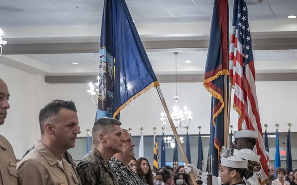 A military ceremony is taking place in an indoor banquet or event hall, featuring a color guard presenting flags including the United States flag and what appears to be a Navy unit flag. Military personnel in various uniforms — including Navy khaki and Marine Corps MARPAT — stand at attention alongside a civilian, while sailors in dress whites carry the flags. An audience of family members and guests is seated and standing in the background.