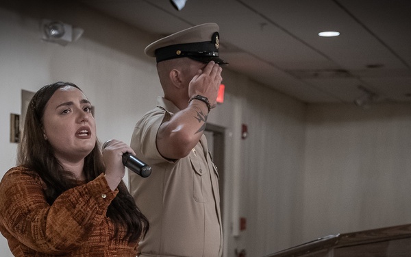 A woman in a rust-colored dress holds a microphone and appears to be singing or speaking at a formal military ceremony. Behind her, a uniformed military officer in tan/khaki dress uniform stands with his hand raised to his face in a salute or emotional gesture. The setting includes a podium and both the American flag and what appears to be the flag of Guam.