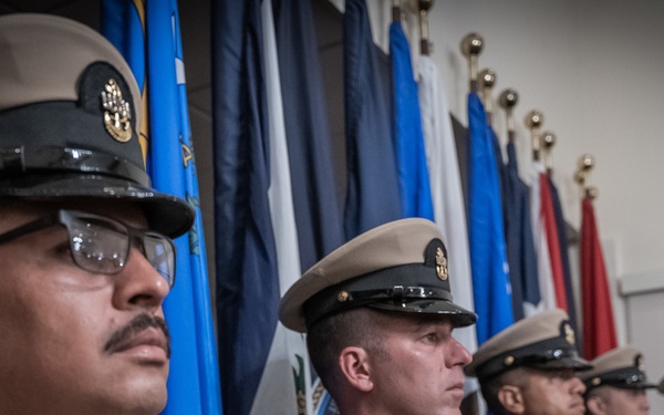 A line of U.S. Navy Chief Petty Officers standing at attention in their khaki service uniforms during what appears to be a formal military ceremony or event. Multiple personnel are aligned in a row against a backdrop of various military and unit flags with gold finials. The foreground subject wears glasses and displays several ribbons and badges on his uniform.