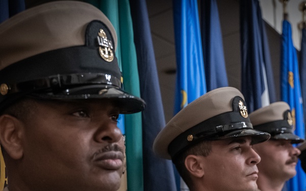 A row of United States Navy Chief Petty Officers stand at attention in their khaki service dress uniforms during what appears to be a formal military ceremony. Multiple service members are aligned in a precise formation against a backdrop of numerous military and organizational flags. The image is captured with a shallow depth of field, emphasizing the foreground sailor while the others recede into soft focus.