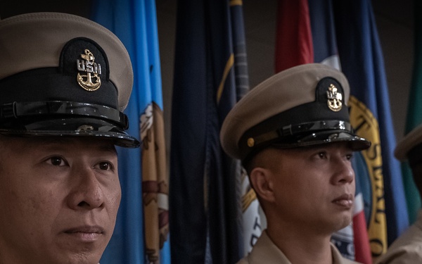 Four U.S. Navy enlisted personnel in Service Khaki uniforms stand at attention in a formal military ceremony setting. They are positioned in a line in front of multiple military and organizational flags. The image has a shallow depth of field with the foreground sailor in sharp focus.