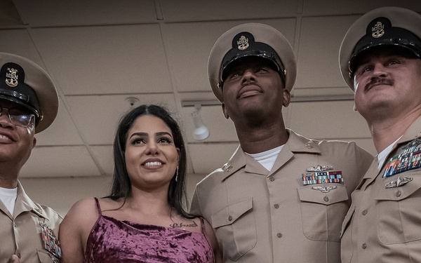 Four individuals pose together for a group photo, three of whom are wearing U.S. Navy Chief Petty Officer uniforms with covers (caps) and service ribbons. The fourth individual is a woman in a burgundy/maroon velvet dress standing between the uniformed personnel. The setting appears to be an indoor military function or ceremony room.