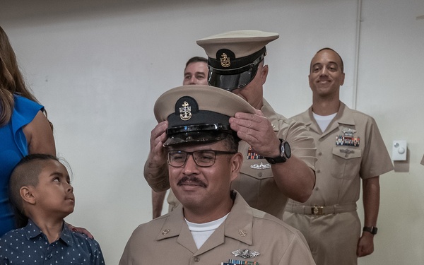 A U.S. Navy chief petty officer promotion ceremony is taking place, with a new chief having his cover (hat) placed on his head by a fellow sailor. The honoree is seated in the foreground wearing a khaki uniform with multiple ribbons and warfare devices, surrounded by family members including a woman in blue, a young girl, and a young boy. Additional Navy personnel in khaki uniforms stand in the background as witnesses to the ceremony.