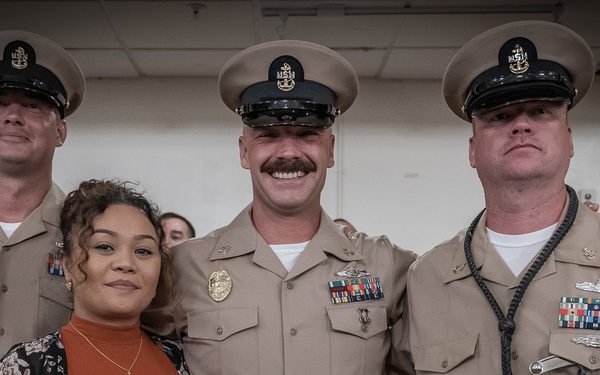 A group photo featuring three U.S. Navy Chief Petty Officers in tan service uniforms and one civilian woman in casual attire. The central figure is smiling broadly and wearing multiple service ribbons and medals, suggesting this may be a promotion or retirement ceremony. The setting appears to be an indoor military facility.
