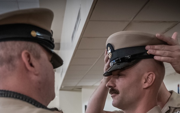 A U.S. Navy promotion or advancement ceremony is taking place indoors, where a senior enlisted sailor is placing a chief petty officer combination cover (hat) on the head of a newly advanced chief petty officer. The recipient is wearing khaki service uniform with multiple ribbons and warfare devices visible on his chest. An audience including civilians and other military personnel is visible in the background.