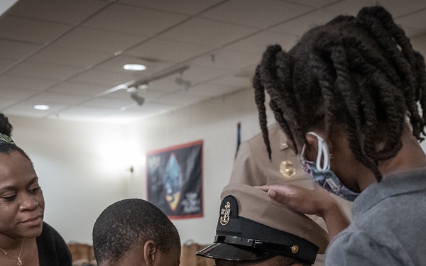 A U.S. Navy service member in dress uniform is being assisted by family members, likely during a promotion or pinning ceremony. Children and a woman are placing or adjusting his cover (hat) and collar devices while he sits or kneels at their level. A second uniformed Navy personnel member stands in the background observing.
