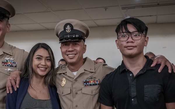 A group photo at what appears to be a U.S. Navy promotion or recognition ceremony. Two uniformed Navy personnel in khaki dress uniforms pose with what appears to be a family group including a woman, two teenage/young adult males, and a young child. The setting is an indoor military facility with a U.S. Navy flag visible in the background.