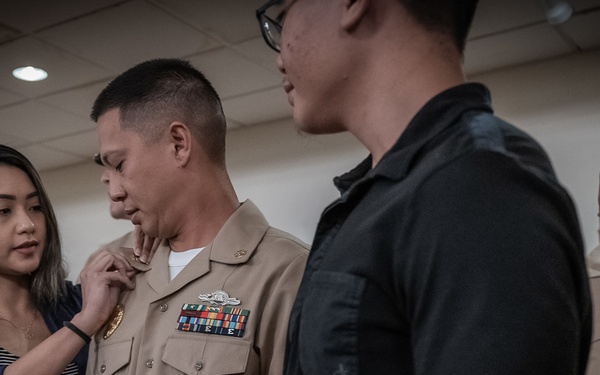 A military promotion or pinning ceremony is taking place, with a woman pinning a new insignia or rank device onto a U.S. Navy sailor in dress khaki uniform. The sailor is surrounded by what appears to be family members, including a young child and a teenager or young adult. A second Navy sailor in uniform is visible in the background observing the ceremony.