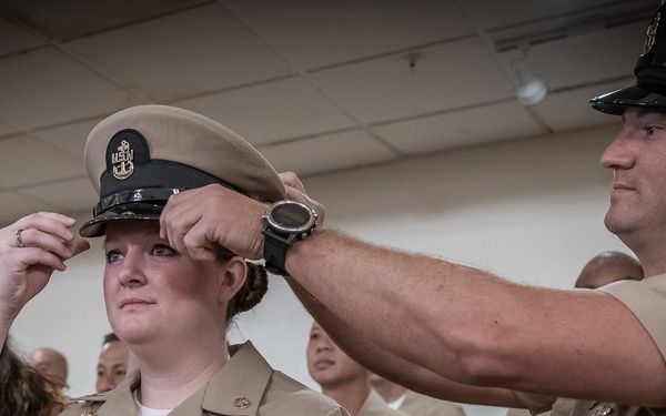 A U.S. Navy promotion ceremony is taking place indoors, where a female sailor is having a chief petty officer combination cover (hat) placed on her head by two individuals — a male senior enlisted sailor in uniform and a civilian woman in a yellow dress. The recipient is wearing a tan Navy service uniform adorned with multiple ribbons and a badge, indicating a significant career milestone. This appears to be a chief petty officer pinning/cover ceremony, a traditional Navy advancement ritual.