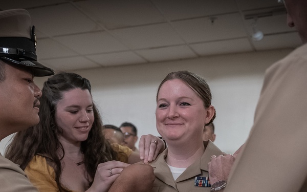 A military promotion ceremony is taking place, with a female sailor in U.S. Navy Service Uniform (tan/khaki) receiving new rank insignia being pinned on both shoulders simultaneously. Two individuals are pinning insignia on her left shoulder — a uniformed male sailor and a civilian woman in a yellow dress — while another uniformed individual (back to camera) pins the right shoulder. The subject is smiling, suggesting this is a celebratory milestone event.