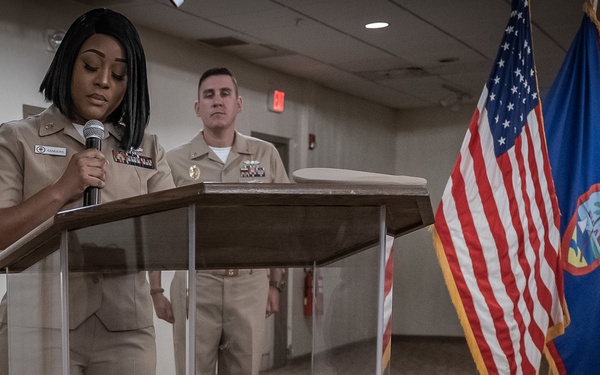 A military ceremony is taking place indoors, with a female Navy sailor speaking at a clear podium while holding a microphone. A male Navy officer stands at attention behind her. Multiple flags are displayed in the background, including the United States flag and the flag of Guam.