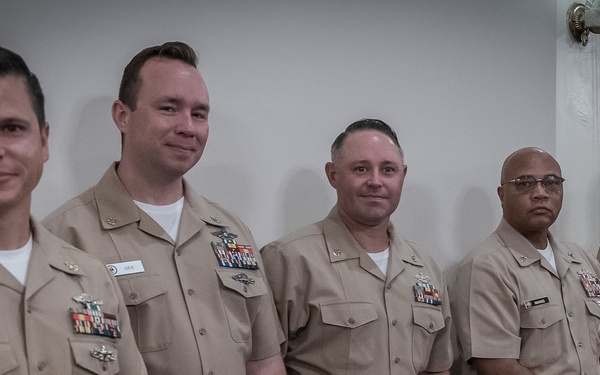 A group of six U.S. Navy personnel in tan/khaki Service Uniform B are posed together, likely at a formal military ceremony or event. Several individuals are holding their combination covers (caps) and display various ribbons, medals, and warfare qualification badges on their uniforms. The setting appears to be an indoor military facility.