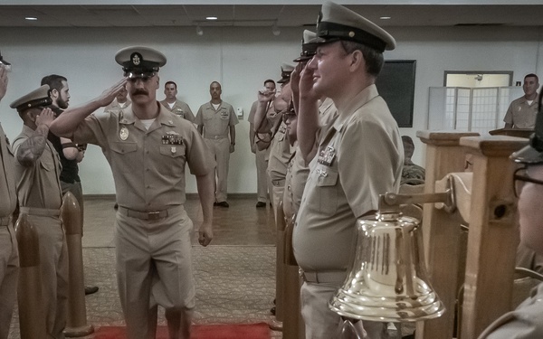 A formal U.S. Navy military ceremony is taking place indoors, with personnel in tan/khaki uniforms lining a red carpet aisle and rendering salutes to an officer walking through. A sailor on the right holds a brass ship's bell, suggesting this is a traditional naval 'side boy' or change of command/retirement ceremony. The scene is well-organized with multiple ranks of personnel standing at attention on both sides.
