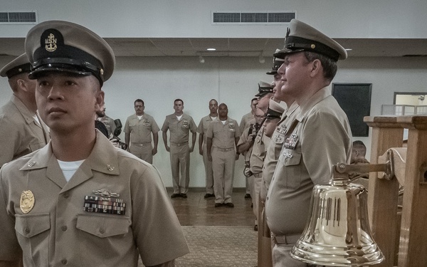 A formal U.S. Navy ceremony is taking place indoors, with personnel in Service Khaki uniforms standing in formation along a red carpet aisle. A brass ship's bell is prominently displayed on a wooden stand, suggesting this is a Chief Petty Officer pinning ceremony or similar naval tradition. Multiple sailors of various ranks are present, with some standing at attention and others observing.