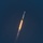 SLD 45 supports the first National Security Space Launch mission on a Falcon Heavy rocket A large rocket is captured mid-flight ascending diagonally through a dark blue sky. Multiple engine plumes produce bright orange and yellow flames with a white exhaust trail extending downward. The rocket's white body and clustered booster configuration are clearly visible against the dark background.