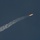 SLD 45 supports the first National Security Space Launch mission on a Falcon Heavy rocket A rocket is captured mid-flight ascending through a dark blue sky, leaving a broad white exhaust plume trail behind it. The rocket is visible in the upper right portion of the image with a bright orange-yellow flame emanating from its engines. The contrail widens as it extends toward the lower left, indicating the rocket has been in flight for a sustained period.
