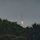 SLD 45 supports the first National Security Space Launch mission on a Falcon Heavy rocket A rocket booster is captured in the process of a vertical landing, with landing legs deployed and a bright engine flame visible just above the treeline. The image is taken at dusk or dawn from a distance, with dense vegetation in the foreground partially obscuring the lower portion of the rocket. A condensation or exhaust trail is visible extending upward from the vehicle into the overcast sky.