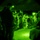 TACP Lightning Challenge 2022 The image is captured through a night vision device, showing a group of military personnel gathered in a dark outdoor environment. Multiple soldiers in combat gear and tactical equipment are visible, some standing and some crouching or seated. A vehicle is partially visible in the background on the right side.