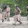 TACP Lightning Challenge 2022 A group of five individuals in tactical/military gear are moving across an open rocky terrain carrying tan-colored jerry cans or water containers. The personnel are wearing a mix of multicam camouflage uniforms and black tactical clothing, with plate carriers, hearing protection, and helmets visible. The setting appears to be an outdoor training exercise or operational activity near a tree line.