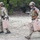TACP Lightning Challenge 2022 Two military personnel in MultiCam/OCP camouflage uniforms and tactical gear are carrying tan-colored jerry cans across an open, rocky terrain. Both individuals are wearing combat helmets and plate carrier vests with visible patches, including what appear to be US and Texas state flag patches. The setting appears to be a training exercise or field operation in a semi-arid environment with sparse vegetation.