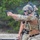 TACP Lightning Challenge 2022 A military or law enforcement operator in full tactical gear is firing a semi-automatic pistol at a shooting range, with a spent casing visibly ejecting from the firearm. The individual is wearing MultiCam camouflage, a tactical helmet with communications equipment, ballistic eyewear, and a plate carrier vest with an American flag patch. A second person in tactical gear is visible in the background, suggesting a supervised training exercise.