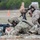 TACP Lightning Challenge 2022 Two soldiers in MultiCam/OCP camouflage uniforms are conducting a marksmanship training exercise at an outdoor firing range. One soldier is in the prone position aiming an M4-style carbine downrange, while a second soldier acts as a spotter using a Leupold spotting scope mounted on a tripod. A humanoid silhouette target is visible in the background at distance.