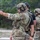 TACP Lightning Challenge 2022 A military or law enforcement personnel in full tactical gear is firing a semi-automatic pistol at a shooting range, with a spent brass casing visibly ejecting from the firearm. Two additional personnel in tactical equipment are visible in the background, one wearing a black shirt with partial text reading 'TOR' suggesting an instructor role. The setting appears to be an outdoor live-fire training range.