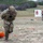 TACP Lightning Challenge 2022 Military personnel are conducting a combat casualty evacuation training exercise at an outdoor range. One fully-equipped soldier in multicam uniform and helmet is dragging an orange rescue litter or training mannequin across open terrain, while a second soldier in multicam without helmet observes and appears to be timing or evaluating the exercise. A shooting target board is visible in the background where additional personnel are present.