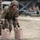 TACP Lightning Challenge 2022 A military personnel in full combat gear, including helmet, body armor, and tactical gloves, is leaning forward to handle two tan/beige water jerry cans labeled 'WATER' and 'FTU'. The scene appears to be an outdoor military training exercise or competition, with other uniformed personnel visible in the background. The setting is a dry, open field with various military equipment and structures visible.