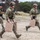 TACP Lightning Challenge 2022 Three personnel in tactical gear are running across an open, arid terrain. Two of the individuals in multicam camouflage uniforms and helmets are each carrying tan/beige jerry cans, while a third person in a black shirt with a tactical vest follows behind. The setting appears to be a training exercise or field operation based on the organized movement and equipment.