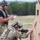 TACP Lightning Challenge 2022 Two military or law enforcement personnel are engaged in a tactical firearms training exercise at an outdoor shooting range. One individual in full combat gear including helmet and plate carrier is in a kneeling position aiming a carbine rifle through an opening in a wooden barricade. A second individual, acting as an observer or evaluator, stands behind holding a clipboard and taking notes while wearing hearing protection and a tactical vest.