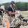 TACP Lightning Challenge 2022 The image depicts what appears to be a military or law enforcement training exercise at an outdoor shooting range. Personnel in tactical gear are practicing shooting techniques, with one individual in full combat kit taking a kneeling position behind a wooden barrier while an observer/instructor with a clipboard monitors and evaluates performance. Additional participants are visible in the background engaging targets.