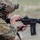 TACP Lightning Challenge 2022 A soldier in full tactical gear is handling an AR-platform rifle, appearing to clear a malfunction or perform a press check near a wooden barrier. The individual is wearing MultiCam camouflage uniform and a tactical helmet equipped with communications gear and accessories. A spent or ejected cartridge casing is visible near the lower receiver of the weapon.
