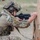 TACP Lightning Challenge 2022 A soldier in full tactical gear is firing a short-barreled AR-platform rifle from a kneeling position behind a wooden barrier. A spent brass casing is visibly ejecting from the weapon, indicating active fire. The setting appears to be a military training range based on the controlled environment and presence of an observer in the background.