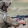 TACP Lightning Challenge 2022 A military or law enforcement operator in full tactical gear is positioned behind a wooden barrier, holding an AR-15/M4-style carbine in a low-ready or reloading position. The individual is wearing MultiCam camouflage, a ballistic helmet with communications equipment, hearing protection, and a plate carrier vest. The setting appears to be an outdoor training range or tactical exercise.