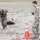 TACP Lightning Challenge 2022 Two soldiers in MultiCam/OCP camouflage uniforms are engaged in what appears to be a combat casualty evacuation (CASEVAC) training exercise on an open field. One fully-equipped soldier wearing a helmet and body armor is dragging a training mannequin/dummy across the ground using a strap or drag handle. A second soldier, acting as an evaluator or observer, stands nearby holding what appears to be a stopwatch or scoring device.